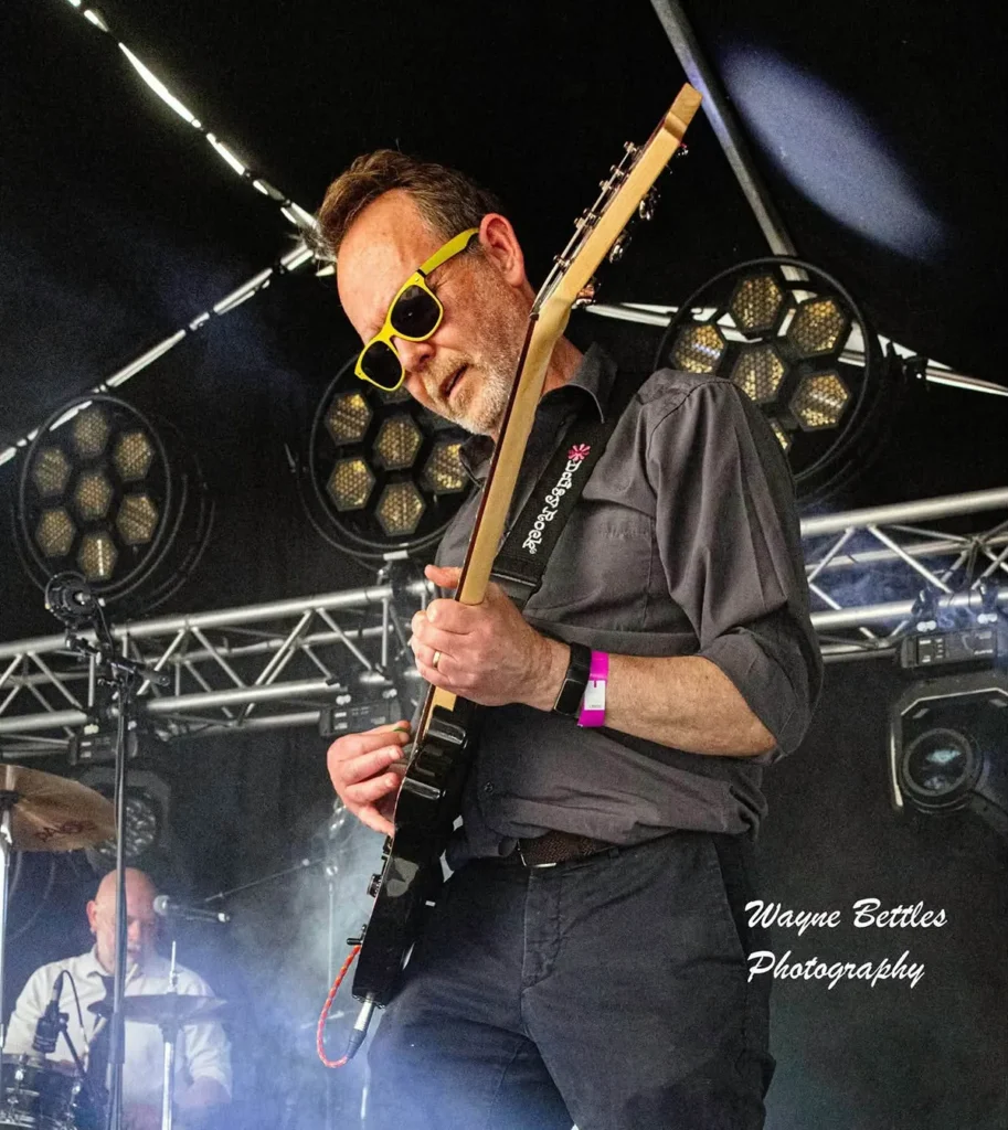 The Expletives performing live on an outdoor stage, guitarist mid-riff under stage lighting